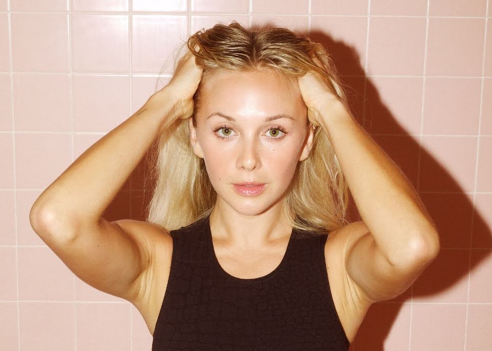 Woman holding her hair back while posing against a soft pink tiled background.