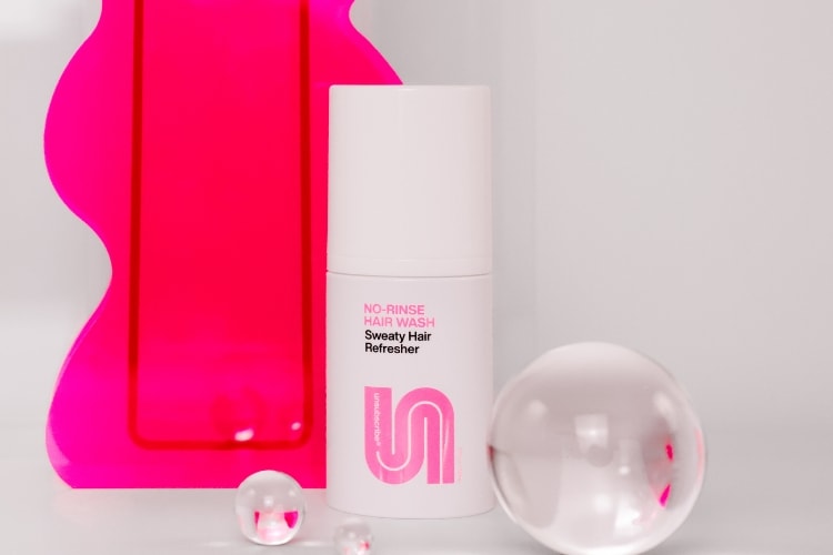 White bottle of No-Rinse Hair Wash Sweaty Hair Refresher placed beside bright pink acrylic decor and clear glass spheres.