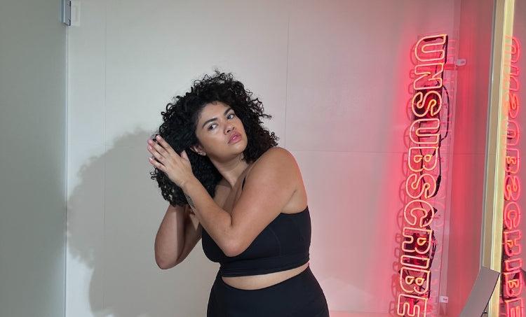 Woman with curly hair adjusting her curls while standing indoors beside a neon “UNSUBSCRIBE” sign.