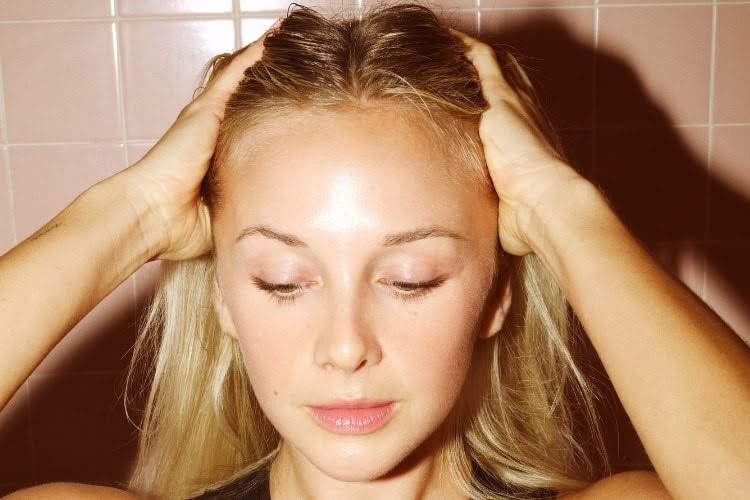 Hand applying white hair product along the scalp and roots of a woman with wet wavy hair.