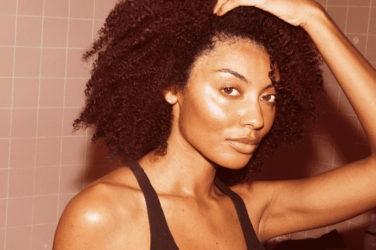 Woman with natural curly hair wearing a black tank top, lifting one hand to her head while looking confidently at the camera against a tiled background.