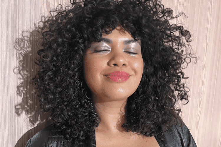 Woman with voluminous curly hair smiling with eyes closed, wearing silver eyeshadow and red lipstick.