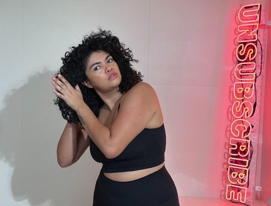 Woman styling her curly hair indoors beside a glowing neon sign.